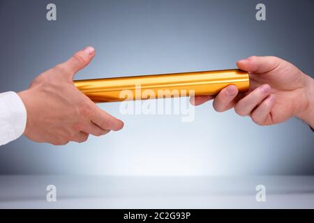 Close-up Of A Hand Passing Golden Relay Baton To Businessman Against Gray Background Stock Photo