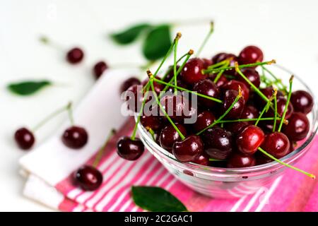 Fresh sour cherries in glass bowl, dishcloth and green leaves on white wooden table. Fresh ripe sour cherries. Stock Photo