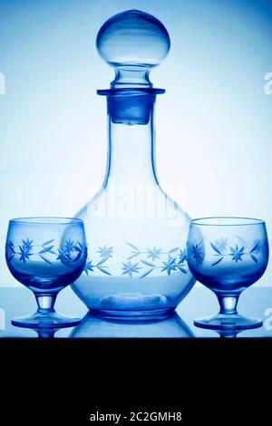 Empty crystal alcoholdecanter and two glasses for vodka standing on a glass table. Vertical side view, cold tone. Stock Photo