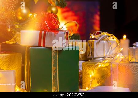 Stack of packed gift boxes under Christmas tree against burning fireplace. Lots of Christmas gifts under the tree. Candles on wooden floor. Focus on g Stock Photo