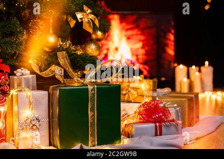 Stack of packed gift boxes under Christmas tree against burning fireplace. Lots of Christmas gifts under the tree. Candles on wooden floor. Focus on g Stock Photo
