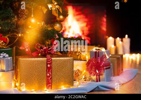 Stack of packed gift boxes under Christmas tree against burning fireplace. Lots of Christmas gifts under the tree. Candles on wooden floor. Focus on g Stock Photo