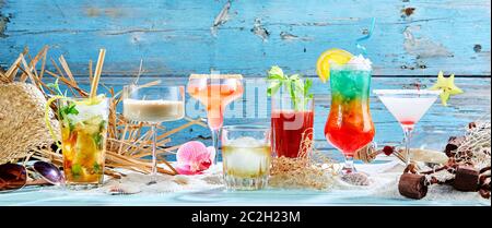 Large variety of tropical cocktails, martini and whisky in a panorama banner arranged on white sea sand over a rustic blue wood background with copy s Stock Photo