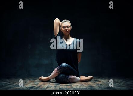 Young woman practicing yoga doing Gomukhasana, cow face pose, in dark room Stock Photo