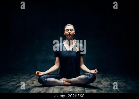 Young attractive woman practicing yoga sitting in lotus pose and meditating in dark room