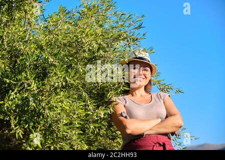Mature beautiful woman in gloves wearing apron hat posing near olive trees Stock Photo