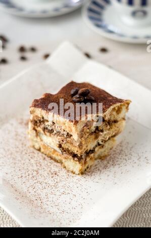 Delicious homemade tiramisu cake slice on a white plate. A classic Italian dessert. In the background espresso coffee cups and spilled coffee beans. Stock Photo