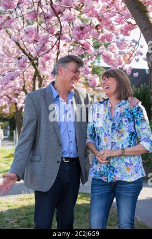 Fun loving senior couple enjoying a joke standing together under a tree with pink spring blossom laughing together. Stock Photo