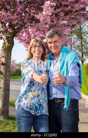 Positive happy senior couple giving a thumbs up as they stand arm in arm under a tree in the street covered in pink spring blossom smiling at the came Stock Photo