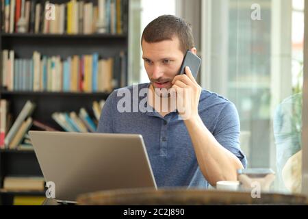 Serious man talks on phone and uses a laptop sitting at home or coffee shop Stock Photo