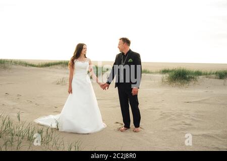 Wedding couple standing and posing on sandy beach. Bride and groom looking to each other and smiling. Barefoot husband. Wedding day concept. Stock Photo