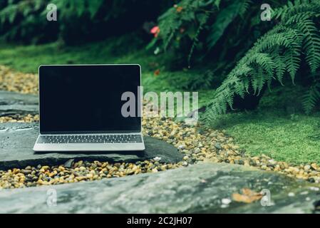 Laptop computer mockup has empty screen outside on stone in garden of coffee shop. Stock Photo