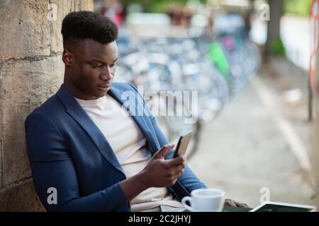 Businessman sitting down at an outdoor table in a city while typing on his mobile phone. Stock Photo