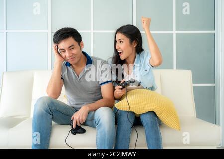 Asian couple man ans woman to playing video games with joysticks while sitting in sofa in living room at home and woman winning in game looking man lo Stock Photo
