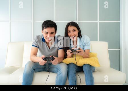 Asian couple man ans woman to playing video games with joysticks while sitting in sofa in living room at home, concept of family values and the modern Stock Photo
