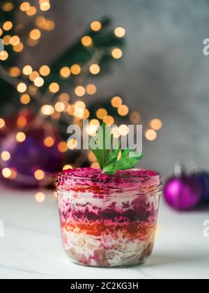 Layered salad herring under a fur coat on festive table, with fir tree and light garland. Traditional russian salad with herring and vegetables in gla Stock Photo