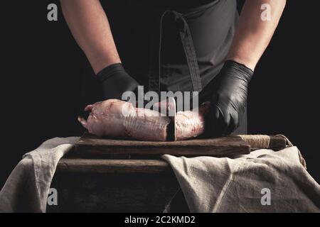 chef in black latex chunks cuts into pieces raw rabbit meat on a brown wooden board, cooking on a dark background Stock Photo