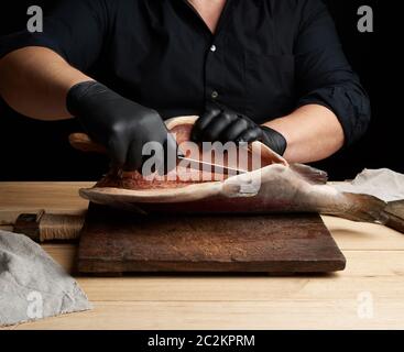 chef in black uniform and black latex gloves cuts a carcass of fresh salmon fish on a wooden cutting board, black background Stock Photo
