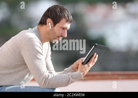 Serious man watching media with tablet and wireless earbuds sitting in a rural town Stock Photo