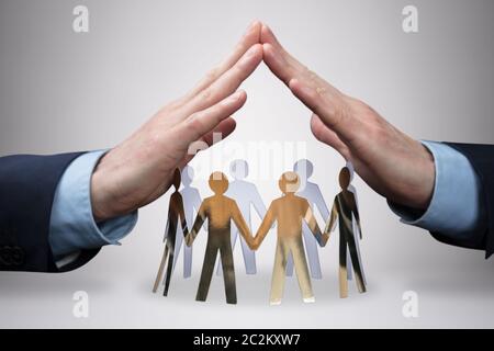 Close-up Of Businessman's Hand Protecting The Circle Of White Paper Cut-out Figures On Gray Background Stock Photo
