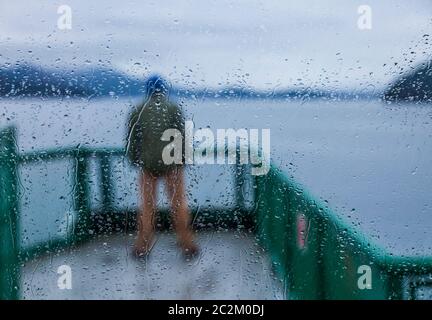 Raindrops cover the windows on a Washington State Ferry and a man stands outside on the observation deck observing the scenery, San Juan Islands, Wash Stock Photo