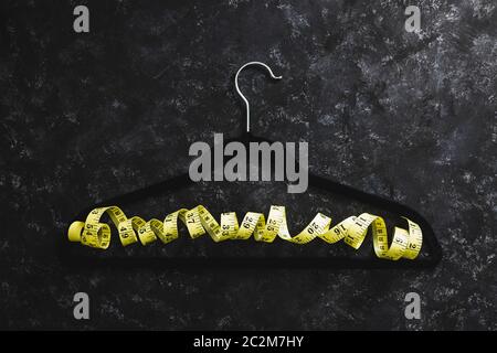 fashion industry and risks of eating disorders concept, velvet hanger with yellow measuring tape on it on black background Stock Photo