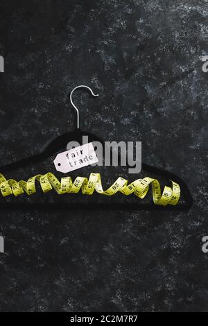 fashion industry and ethical products concept, velvet hanger with yellow measuring tape and Fair Trade label on it on black background Stock Photo