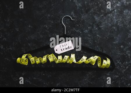 fashion industry and ethical products concept, velvet hanger with yellow measuring tape and Fair Trade label on it on black background Stock Photo