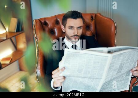 Confident young businessman reading newspaper and latest news while sitting in armchair Stock Photo