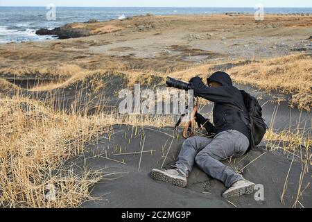 Photographer in Iceland lying in the black sand of a beach using telephoto lens outdoors Stock Photo