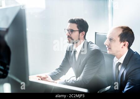 Image of two thoughtful businessmen looking at data on multiple computer screens, solving business issue at business meeting in modern corporate offic Stock Photo