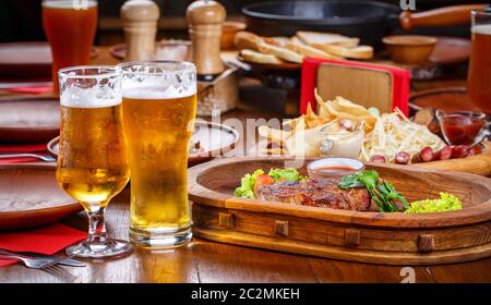 Hot steak with tomatoes, salad, zucchini, greens and sauce on a wooden board. Two glasses of light beer. Beer table in the pub Stock Photo