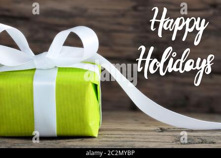 Label With English Calligraphy Happy Holidays. One Green Gift With White Bow. Brown Wooden Background Stock Photo