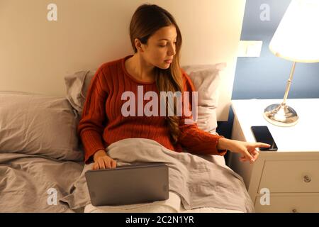 Student girl working on laptop on bed distracted by phone at the night time Stock Photo