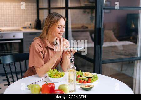 Carefree happy young woman uses smartphone for taking photos of vegetables. Stock Photo