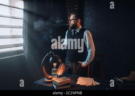 Thoughtful bearded writer in glasses smoking a cigarette and look at the window with sunlight. Retro typewriter, feather, crystal decanter, books and Stock Photo