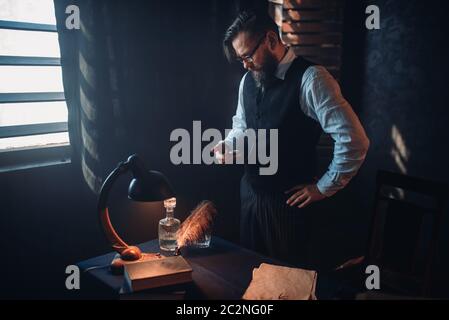 Thoughtful bearded writer in glasses smoking a cigarette and look at the window with sunlight. Retro typewriter, feather, crystal decanter, books and Stock Photo