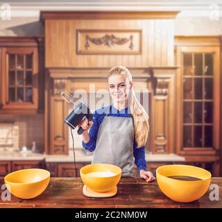Young woman in apron holds mixer in hands against wooden table with bowls for dough. Sweet cake cooking preparation. Kitchen on background Stock Photo