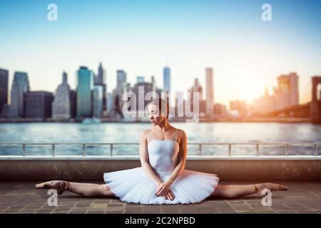 Body flexibility of classical ballet dancer. Ballerina in white dress sits on a twine, front view, cityscape with skyscrapers on background