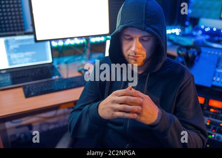 Hacker in mask and hood sitting at his workplace with laptops and PC, information hacking. Internet spy, illegal lifestyle Stock Photo