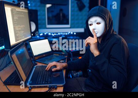 Hacker in mask and hood sitting at his workplace with laptop and PC, network or account hacking. Internet spy, illegal lifestyle, risk job Stock Photo