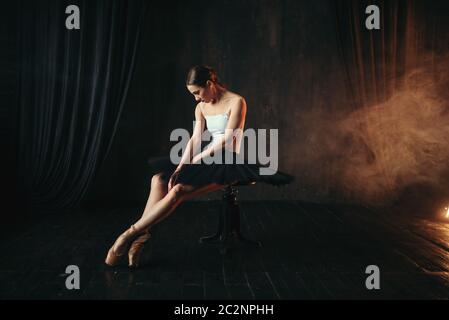 Ballet dancer sitting on black banquette on the stage in theatre. Graceful ballerina training in class Stock Photo