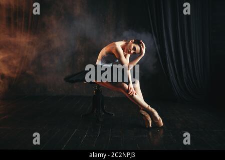 Ballerina sitting on black banquette on the stage in theatre. Elegance ballet dancer poses in class Stock Photo
