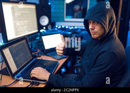 Hacker in hood shows thumbs up at his workplace with laptop and PC, password or account hacking. Internet spy, crime lifestyle, risk job, network crim Stock Photo