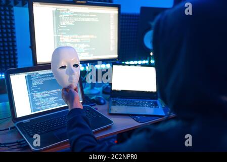 Hacker in hood holds mask in hand and at his workplace with laptop and PC, password or account hacking. Internet spy, crime lifestyle, risk job, netwo Stock Photo