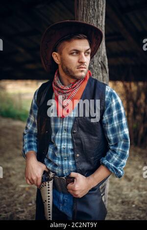 Brutal cowboy in jeans and leather jacket, texas ranch on background, western. Vintage male person with revolver, wild west lifestyle Stock Photo