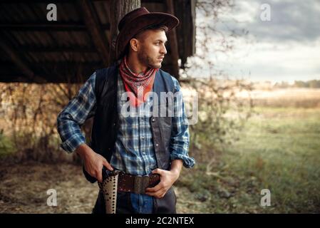 Brutal cowboy with his hand on the revolver, texas ranch on background, western. Vintage male person with gun, wild west lifestyle Stock Photo