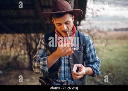 Brutal cowboy lights a cigar with matches, texas ranch on background, western. Vintage male person relax on farm, wild west lifestyle Stock Photo