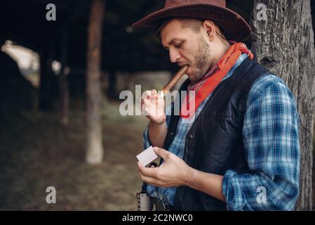 Brutal cowboy lights a cigar with matches, texas ranch on background, western. Vintage male person relax on farm, wild west lifestyle Stock Photo