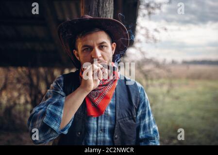 Brutal cowboy smokes a cigar, texas ranch on background, western. Vintage male person with gun on farm, wild west culture Stock Photo